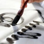 Close-up of a hand using a calligraphy brush to apply black ink on white paper.